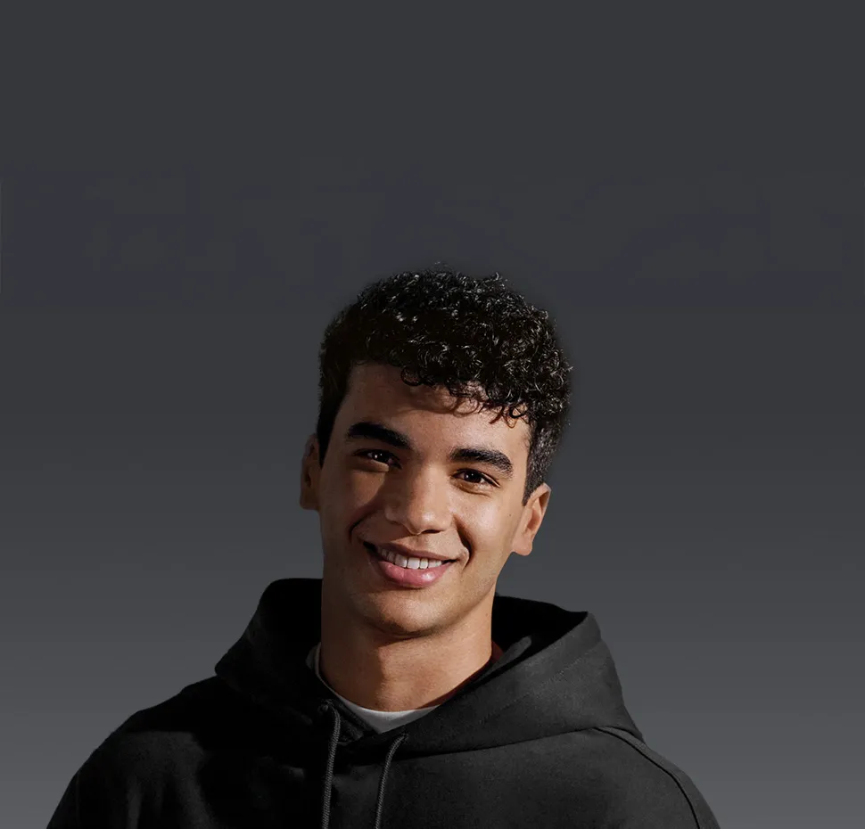 Młody mężczyzna w czarnej bluzie z kapturem uśmiecha się do kamery.