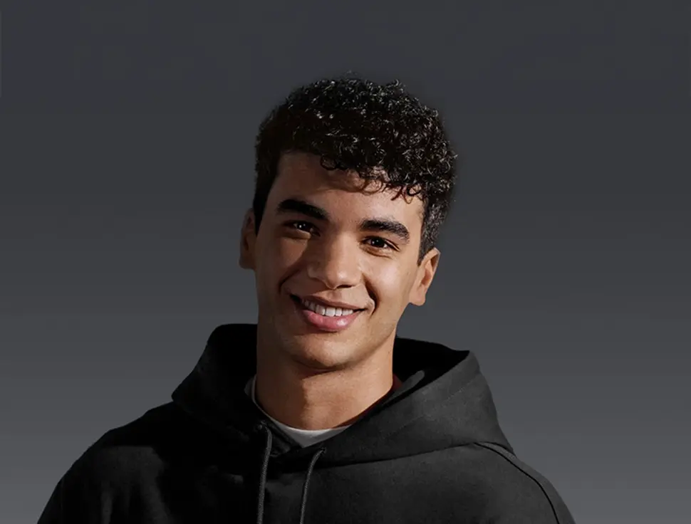 Młody mężczyzna w czarnej bluzie z kapturem uśmiecha się do kamery.