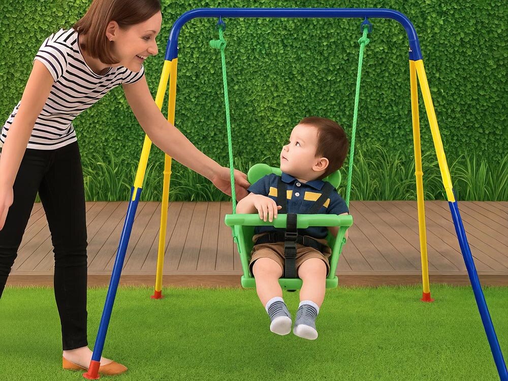 Na obrazie znajduje się dziecko siedzące na zielono-niebieskiej huśtawce, a obok stoi dorosła osoba. W tle widoczna jest zielona ściana z roślinnością i drewniana podłoga.
