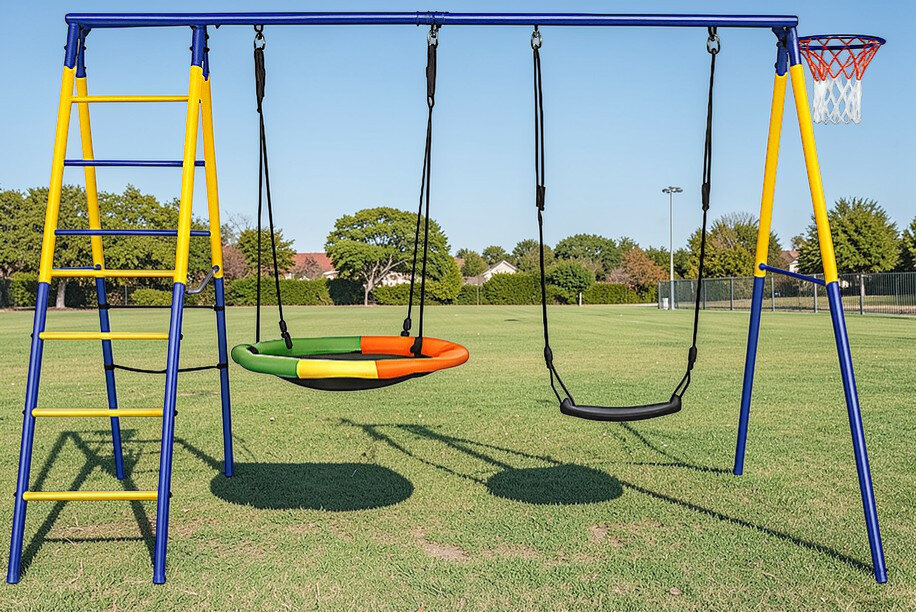 Na trawniku stoi metalowa konstrukcja z dwoma huśtawkami: jedna okrągła w kolorach zielonym, pomarańczowym i żółtym, druga czarna. Obok znajduje się drabinka i kosz do koszykówki.
