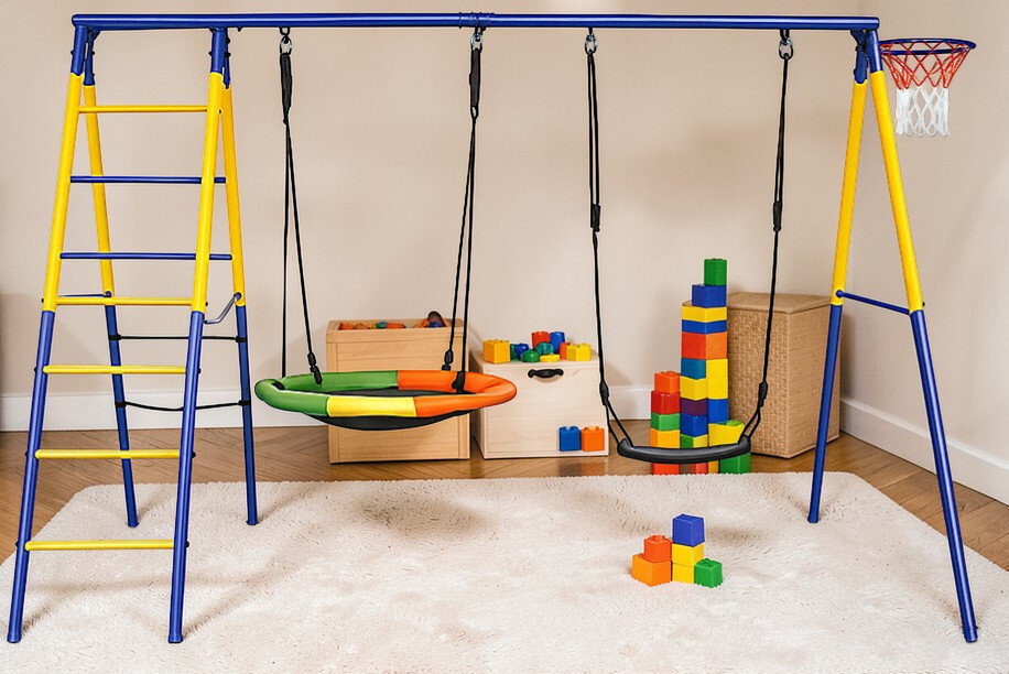 Na obrazie znajduje się kolorowa huśtawka ogrodowa z dwoma siedziskami, umieszczona na metalowej konstrukcji z drabinką i koszem do koszykówki. W tle widoczne są drewniane skrzynie z zabawkami i kolorowe klocki na dywanie.