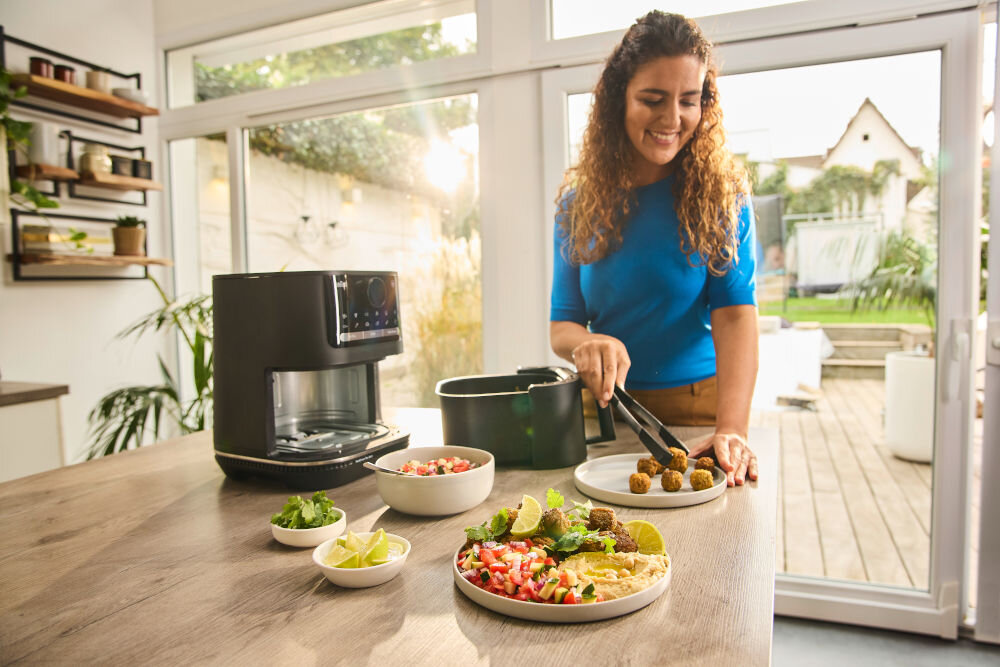 Air Fryer Frytkownica beztłuszczowa BRAUN MultiFry 5 HF5073 uśmiechająca się kobieta frytkownica stoi na blacie kobieta wykłada potrawę na talerz z 20 automatycznymi programami gotowania szybkie łatwe zdrowe gotowanie nie wymaga wstępnego nagrzewania zaawansowane funkcje szeroka gama potraw domowe frytki z różnych warzyw grillowane pieczone mięsa delikatne ryby i owoce morza dodatki do dań głównych przekąski domowe ciasta smaczne zdrowe posiłki