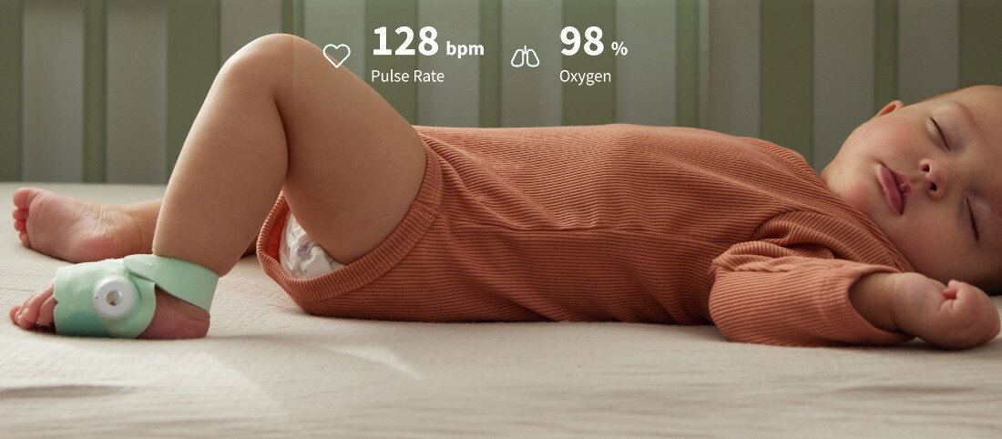 Dziecko leży na boku w brązowym ubranku, na stopie ma założony zielony monitor. Na obrazie widoczny jest tekst: \'128 bpm Pulse Rate\' oraz \'98% Oxygen\'.