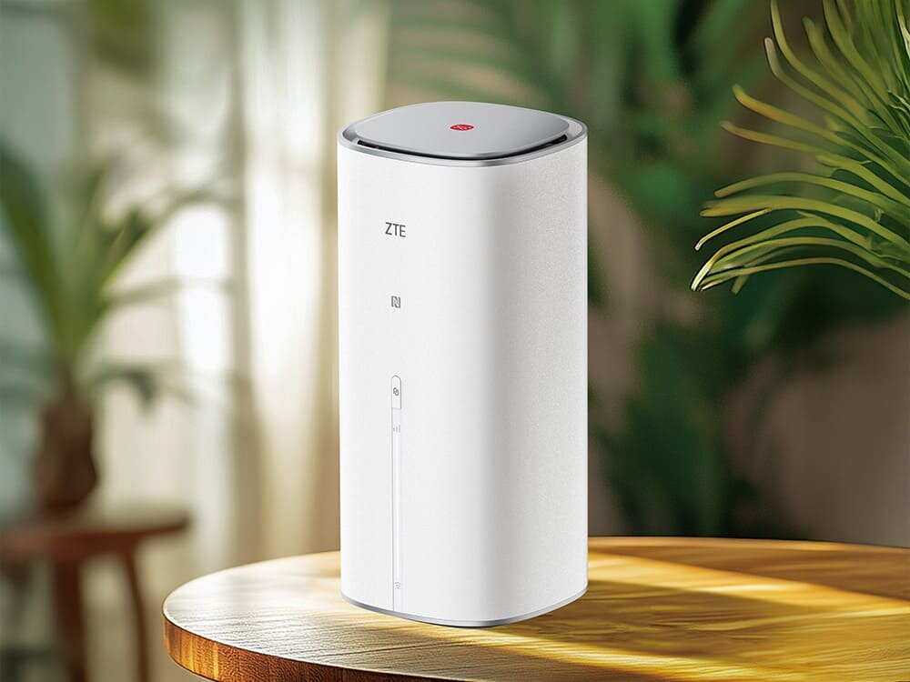 Router ZTE G5 Ultra MC8512 Biały, cylindryczny router ZTE G5 Ultra z czerwonym przyciskiem na górze. Stoi na drewnianym blacie w otoczeniu roślin, symbolizując szybki dostęp do internetu w domowym otoczeniu.