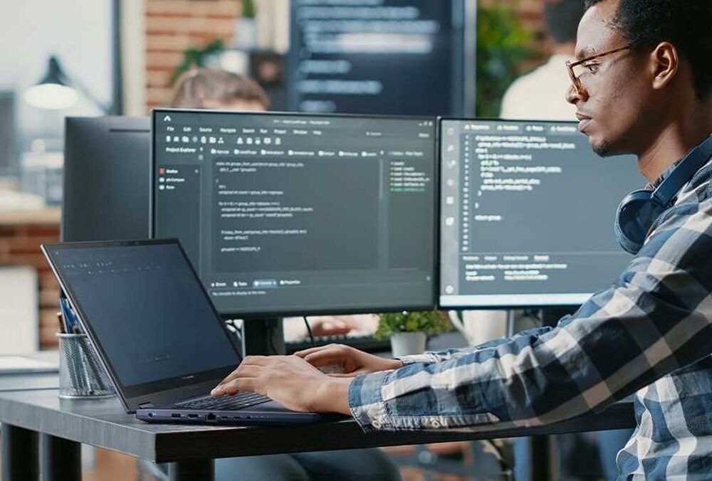 ASUS ExpertBook BM1 z programistą pracującym na laptopie i dwóch zewnętrznych monitorach w nowoczesnym biurze, demonstrując bezpieczeństwo klasy biznesowej dla deweloperów - Bezpieczeństwo klasy biznesowej 