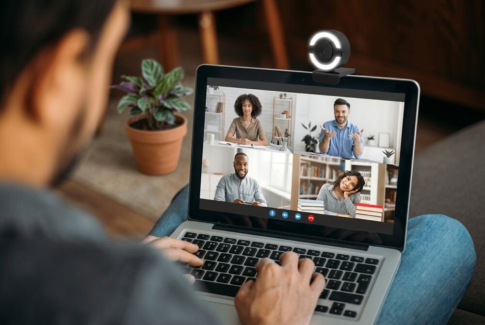 Osoba korzysta z laptopa, na którym odbywa się wideokonferencja z czterema uczestnikami. Na górze laptopa zamontowana jest kamera internetowa z podświetleniem.