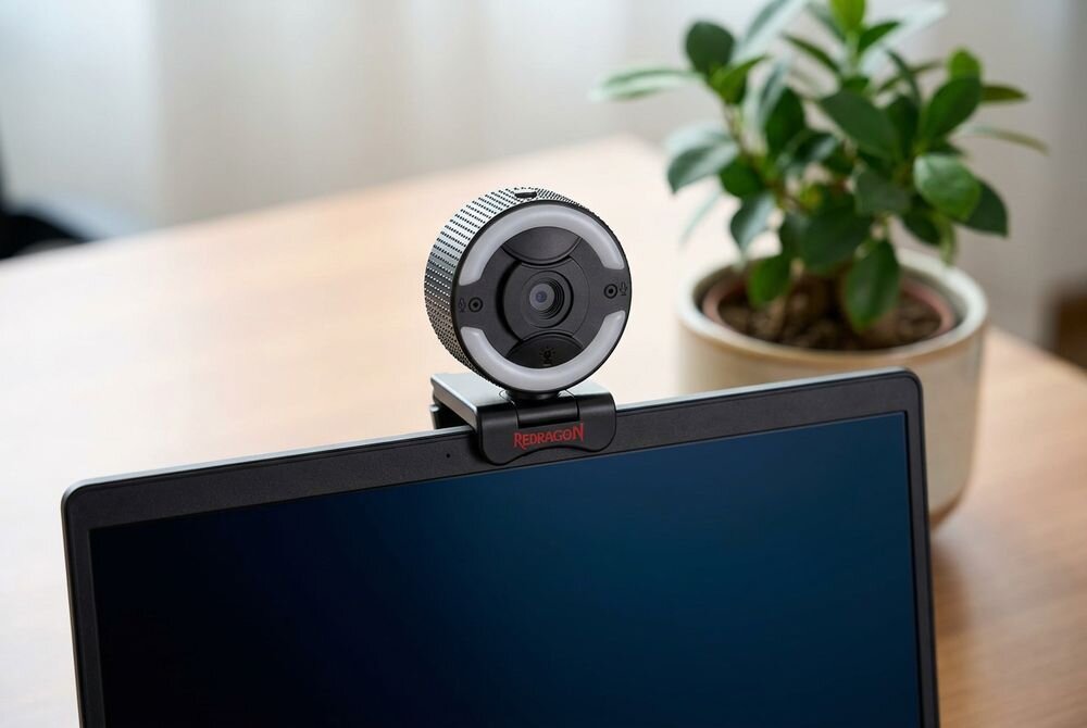 Kamera internetowa zamontowana na górnej krawędzi monitora z widocznym logo \'REDRAGON\'. W tle znajduje się roślina doniczkowa na drewnianym biurku.