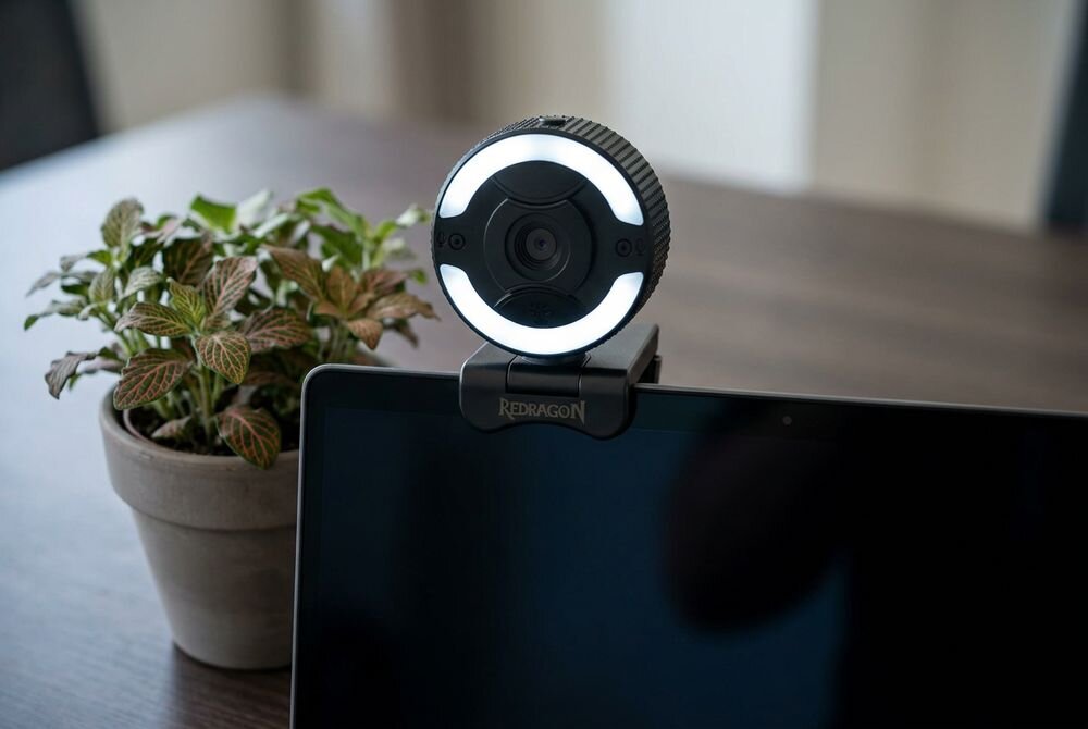 Na obrazie widać kamerę internetową zamontowaną na monitorze, z podświetleniem LED wokół obiektywu. Obok znajduje się doniczka z rośliną, a na uchwycie kamery widoczny jest napis \'REDRAGON\'.