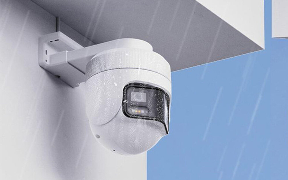 Biała, obrotowa kamera zewnętrzna zamontowana na ścianie, pokryta kroplami deszczu. W tle widoczne jest niebieskie niebo.