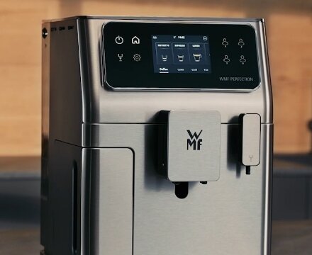Ekspres do kawy z panelem dotykowym wyświetlającym różne opcje napojów, z widocznym logo \'WMF Perfection\'. Urządzenie ma metaliczną obudowę i nowoczesny design.