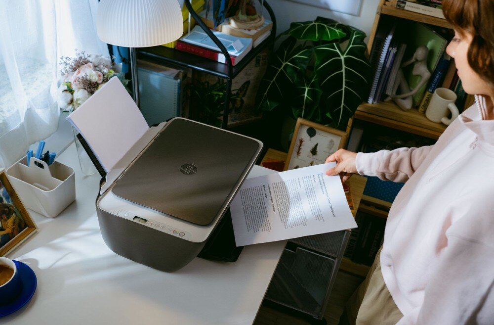 Urządzenie wielofunkcyjne HP DeskJet 2910 Widok z góry na jasne biurko, na którym stoi urządzenie wielofunkcyjne z wysuniętą górną taca podawania papieru. Kobieta prawą ręką odbiera zadrukowaną kartkę papieru z tacy odbiorczej, rozdzielczość, szybkość druku