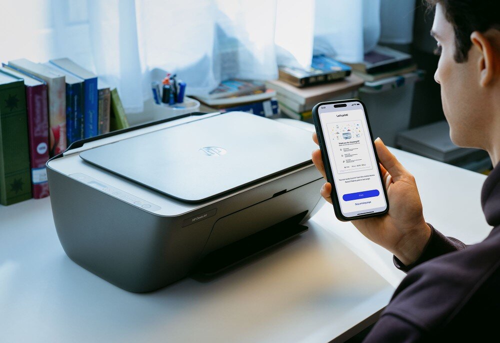 Urządzenie wielofunkcyjne HP DeskJet 2910 Zbliżenie na dłoń mężczyzny trzymającego smartfon z uruchomioną aplikacją mobilną do obsługi druku, na której widnieje przycisk 'Print'. W tle na białym blacie stoi urządzenie wielofunkcyjne ze srebrną górną pokrywą, interfejs, obsługa, aplikacja