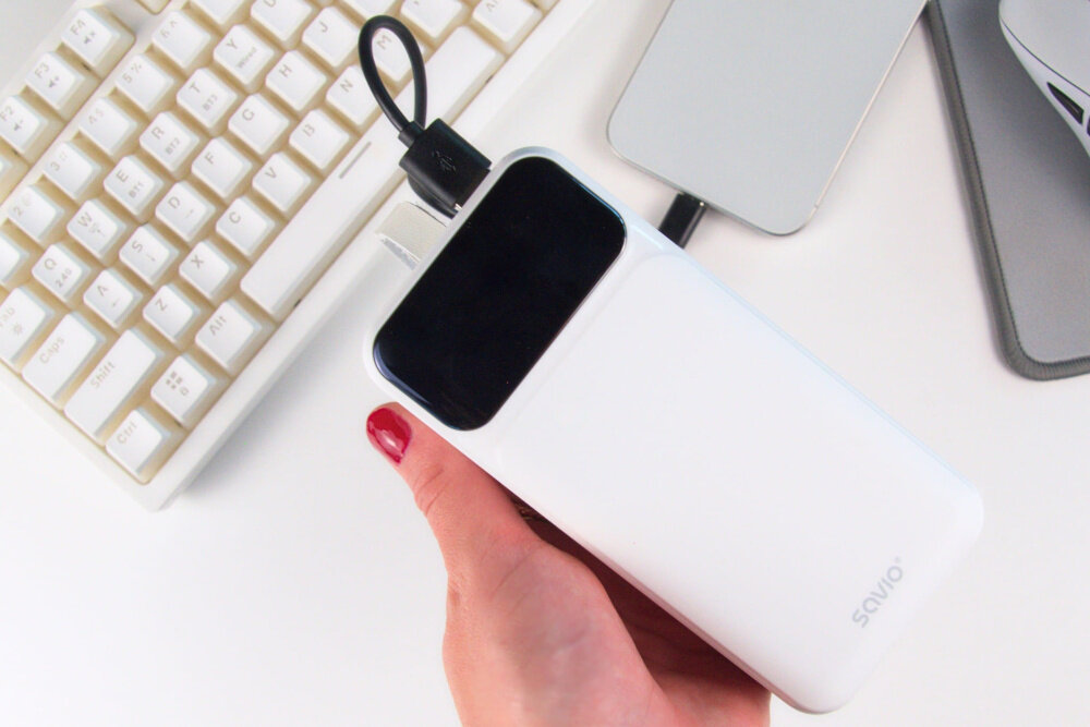 Osoba trzyma biały powerbank z czarnym panelem, podłączony kablem USB. Na urządzeniu widoczne jest logo \'savio\'.