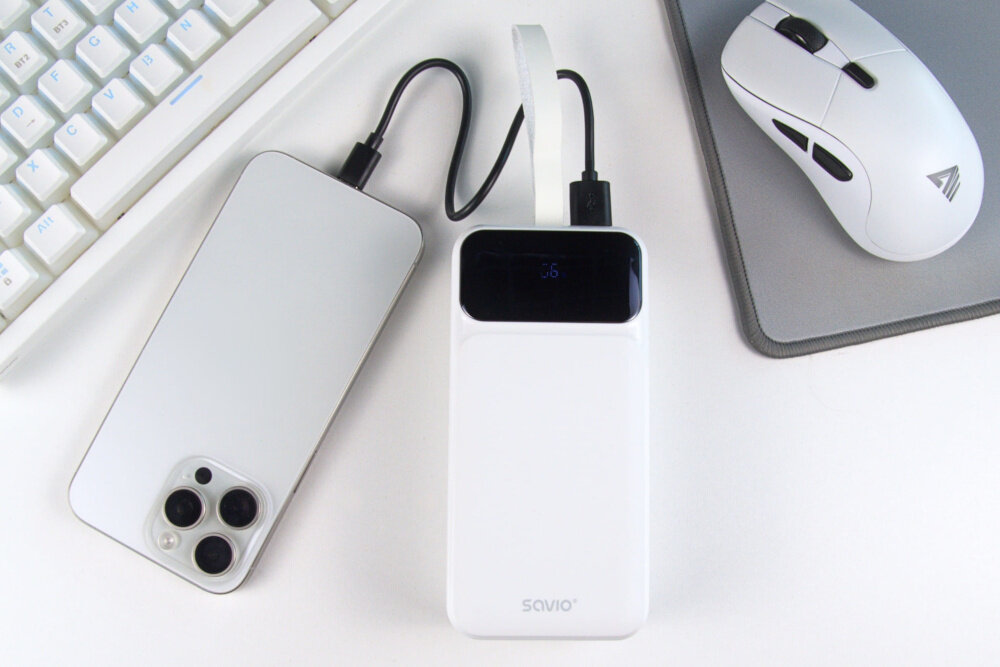 Na białym biurku znajduje się biały powerbank z wyświetlaczem, na którym widoczna jest liczba 76, obok leży smartfon oraz mysz komputerowa. Na powerbanku widnieje napis \'savio\'.