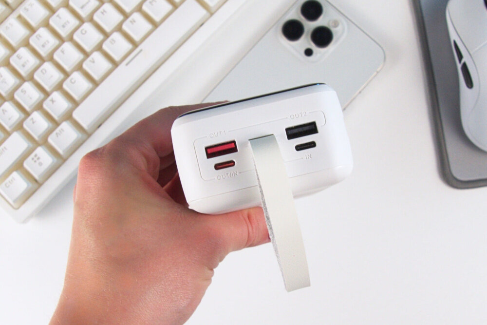 Osoba trzyma biały powerbank z dwoma portami USB oznaczonymi jako \'OUT 1\' i \'OUT 2\'. W tle widoczna jest klawiatura, telefon i myszka.