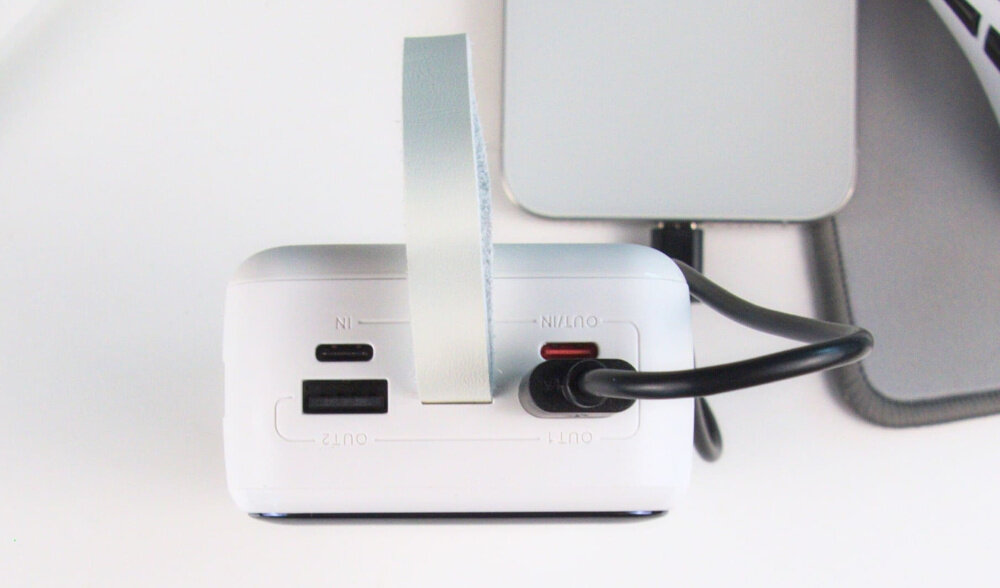 Biały powerbank z dwoma portami USB i jednym portem USB-C, z podłączonym czarnym kablem. Na obudowie widoczne są oznaczenia \'IN\', \'OUT1\', \'OUT2\'.