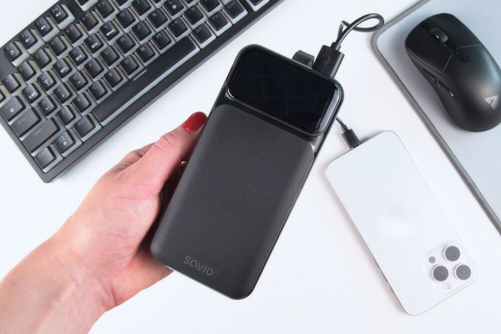 Osoba trzyma czarny powerbank z napisem \'savio\', obok znajduje się klawiatura, myszka i biały smartfon podłączony do powerbanka.