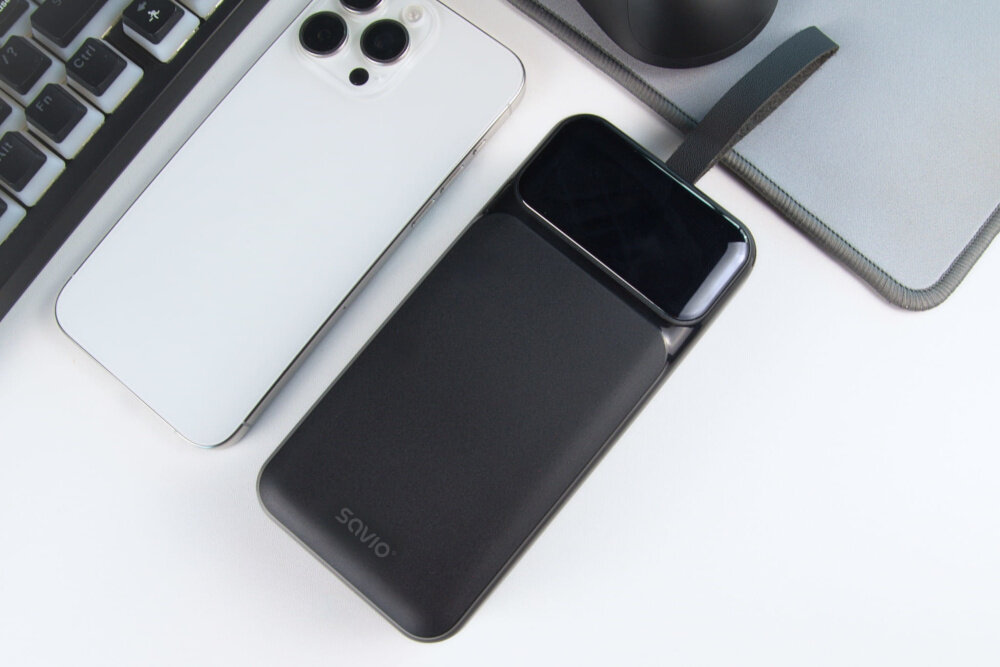 Na obrazie widoczny jest czarny powerbank z logo \'Savio\', obok białego smartfona i klawiatury. Powerbank ma prostokątny kształt i błyszczącą powierzchnię na górze.