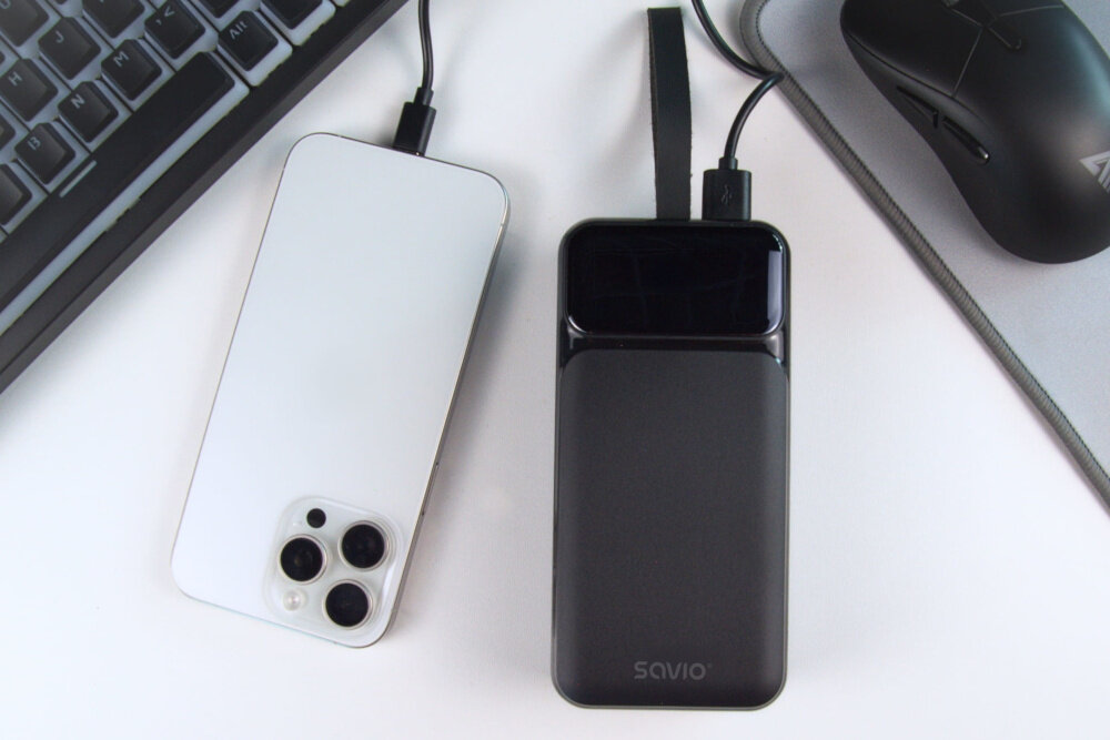 Na białym tle znajduje się czarny powerbank z napisem \'SAVIO\', obok którego leży biały smartfon z trzema obiektywami aparatu. W tle widoczna jest część klawiatury i myszki na podkładce.
