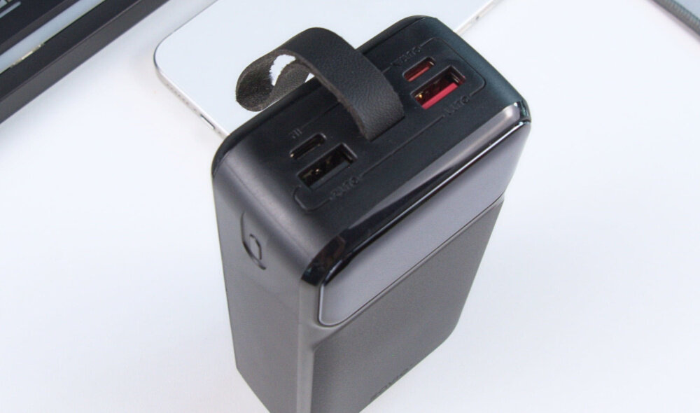 Czarny powerbank z trzema portami USB i skórzanym uchwytem na górze. Obok widoczny fragment białego urządzenia elektronicznego.