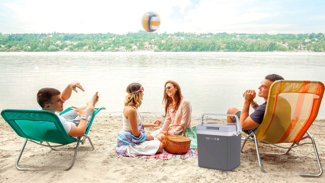 Grupa osób siedzi na plaży przy jeziorze, obok nich znajduje się przenośna lodówka TEESA Easy Cool A25 TSA5001.1. W tle widać zalesiony brzeg jeziora.