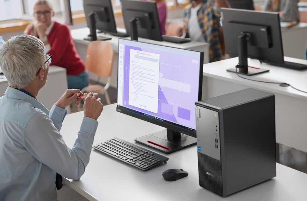 Komputer LENOVO ThinkCentre neo 50t Gen 6 Zdjęcie ukazuje kobietę o siwych włosach siedzącą tyłem do obiektywu przy biurku z komputerem stacjonarnym i monitorem. Na ekranie widać okno aplikacji do kodowania, pamięć RAM, dysk, pojemność