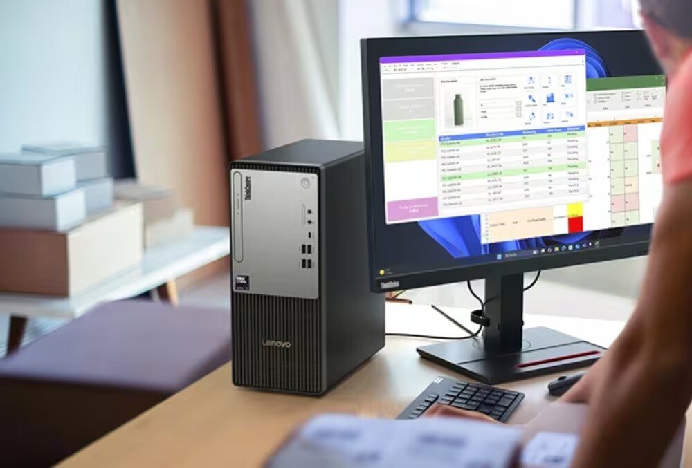 Komputer LENOVO ThinkCentre neo 50t Gen 6 Zdjęcie prezentuje komputer stacjonarny stojący na jasnym, drewnianym biurku obok monitora. Na ekranie widoczna jest aplikacja do zarządzania zamówieniami i logistyką z tabelami i zdjęciem produktu, układ TPM, zabezpieczenie obudowy, chłodzenie powietrzne