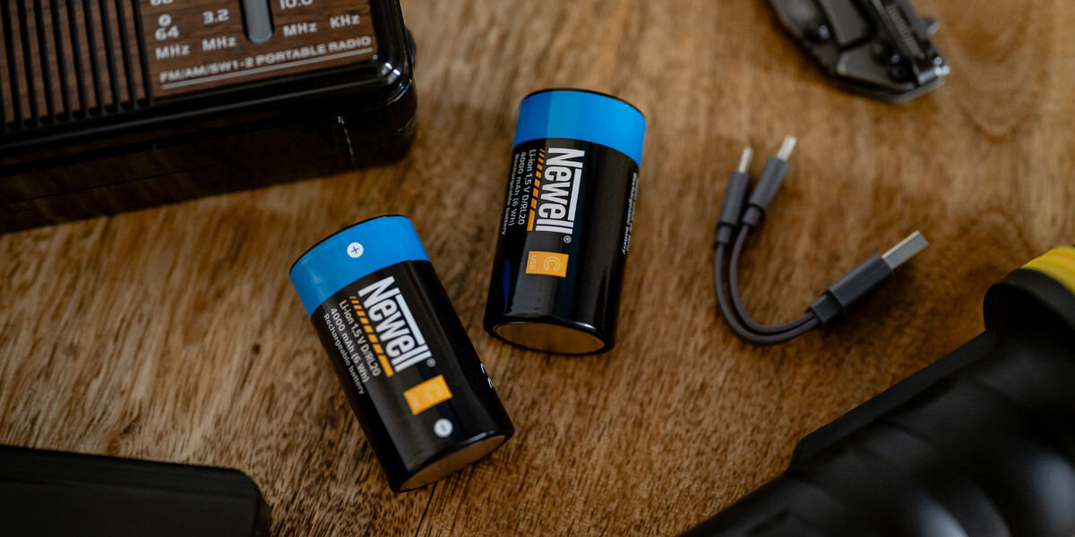 Akumulatorki NEWELL 21700 USB-C 5000 mAh (2 szt.), energia, akumulator, moc, Power bank czarny podłączony kablem USB-C do dwóch akumulatorów Newell USB-C C/R14 na drewnianym stole, obok retro radia i narzędzia, pokazujące ładowanie z power banka bez dedykowanej ładowarki.