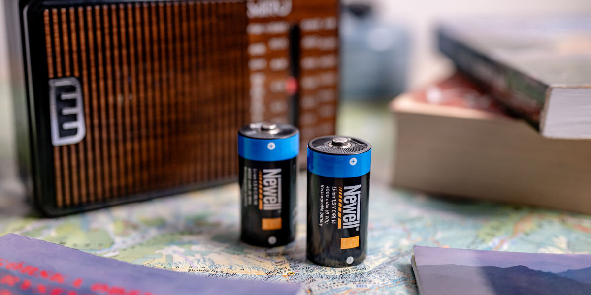 Akumulatorki NEWELL 21700 USB-C 5000 mAh (2 szt.), ogniwa, D, pojemność, zasilanie, Blister z dwoma niebiesko-pomarańczowymi akumulatorami Newell USB-C C/R14 4000 mAh obok retro radia FM z pokrętłami (3V-4.5V), na drewnianym stole
