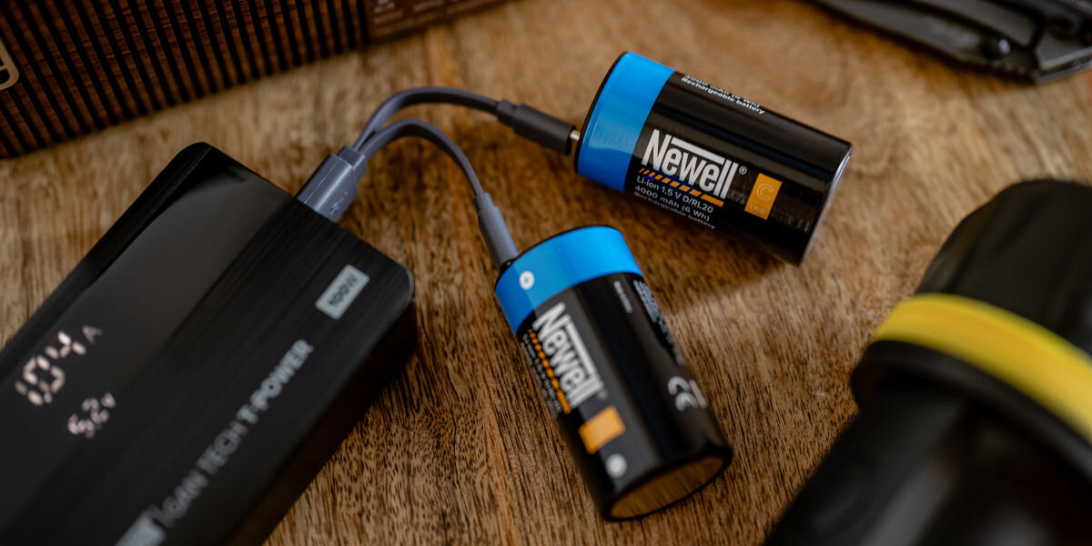 Akumulatorki NEWELL 21700 USB-C 5000 mAh (2 szt.), USB-C, ładowanie, mobilność, wygoda, Dwa akumulatory Newell USB-C C/R14 ładowane kablem USB-C od power banka, obok aparatu i mapy, ilustrujące szybkie doładowanie w podróży dzięki portowi USB-C.