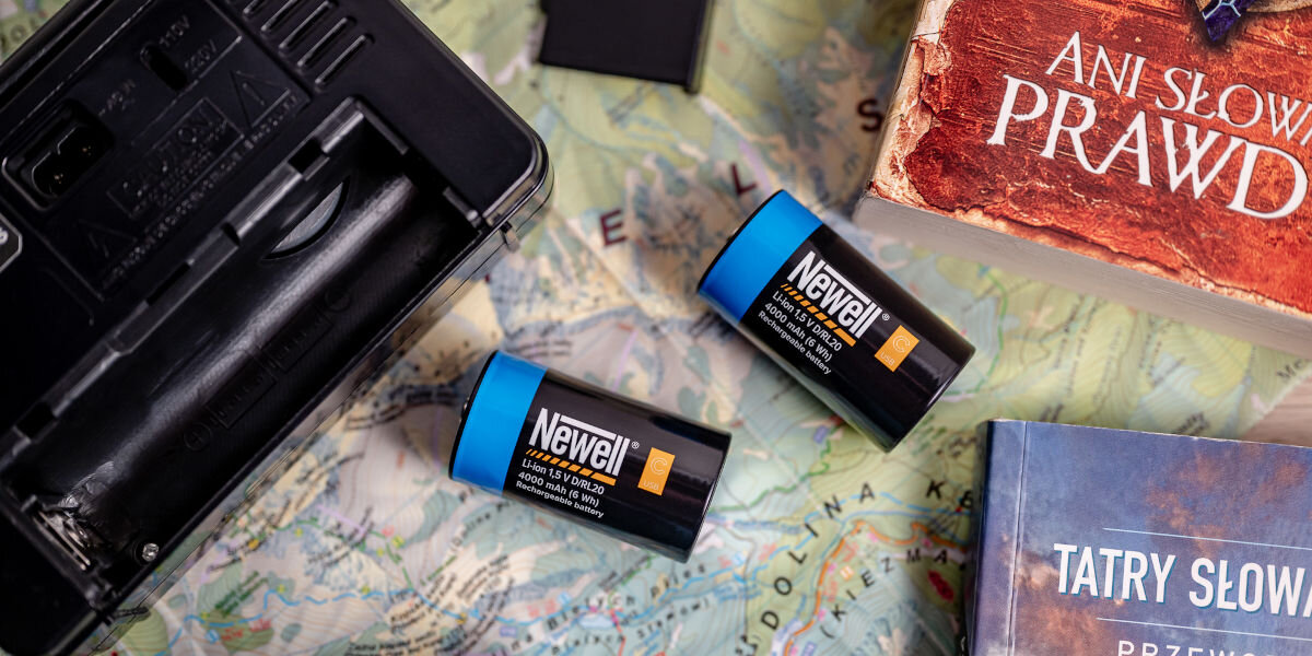 Akumulatorki NEWELL 21700 USB-C 5000 mAh (2 szt.), trwałość, ekologia, oszczędność, wydajność, Dwa akumulatory Newell USB-C C/R14 (niebieski i czarny) obok retro radia, mapy i książek podróżniczych na drewnianym stole, podkreślające uniwersalność do sprzętu audio i gier planszowych.