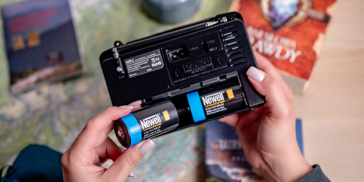 Akumulatorki NEWELL 21700 USB-C 5000 mAh (2 szt.), bezpieczeństwo, ochrona, elektronika, stabilność, Dłoń w szarym swetrze wkłada dwa akumulatory Newell USB-C C/R14 (niebieski i pomarańczowy) do baterii radia przenośnego, obok mapy podróży i książki