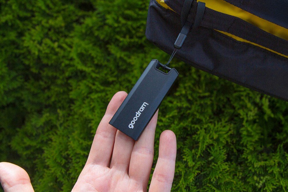 Dysk GOODRAM Move Ridge Pendrive z białym napisem 'goodram' wisi na smyczy nad otwartą dłonią, w tle zielone krzewy i fragment torby, mobilność, wymiary, obudowa