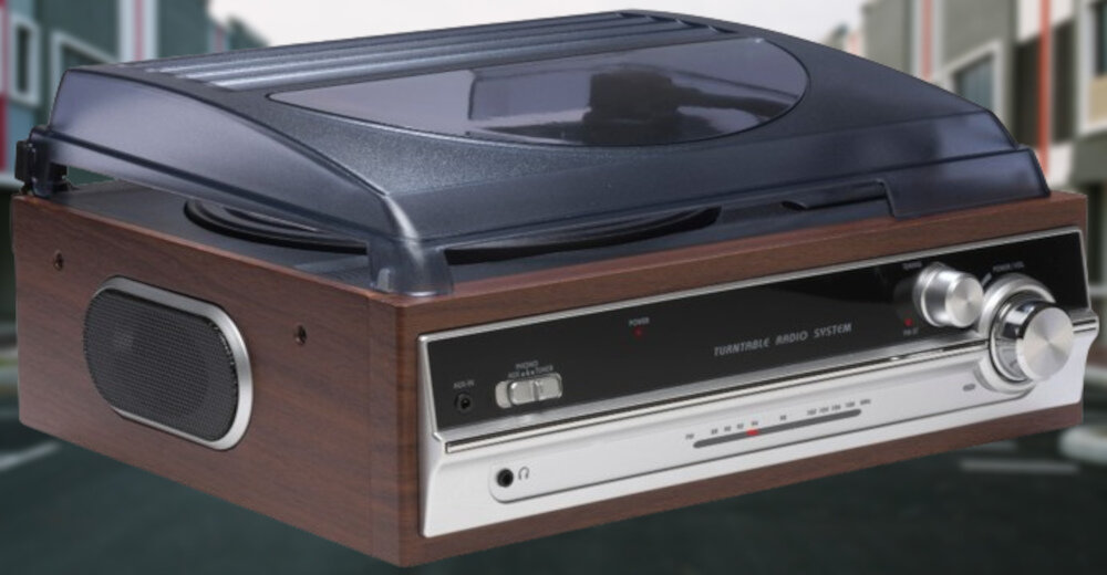 Gramofon w kolorze orzechowym z przezroczystą pokrywą i wbudowanymi głośnikami. Na panelu przednim widoczne są pokrętła i przełączniki oraz napis \'TURNTABLE AUDIO SYSTEM\'.