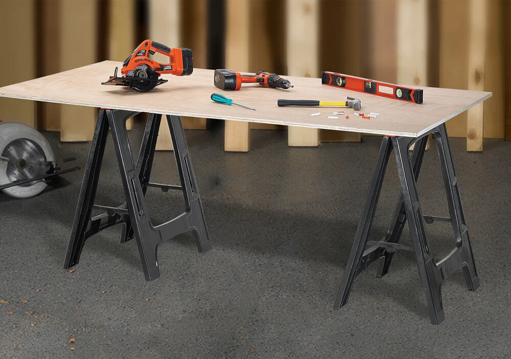 Na obrazie widoczny jest stół roboczy z blatem podpartym na dwóch czarnych kobyłkach warsztatowych. Na blacie znajdują się narzędzia, takie jak piła tarczowa, wkrętarka, poziomica, młotek i śrubokręt.