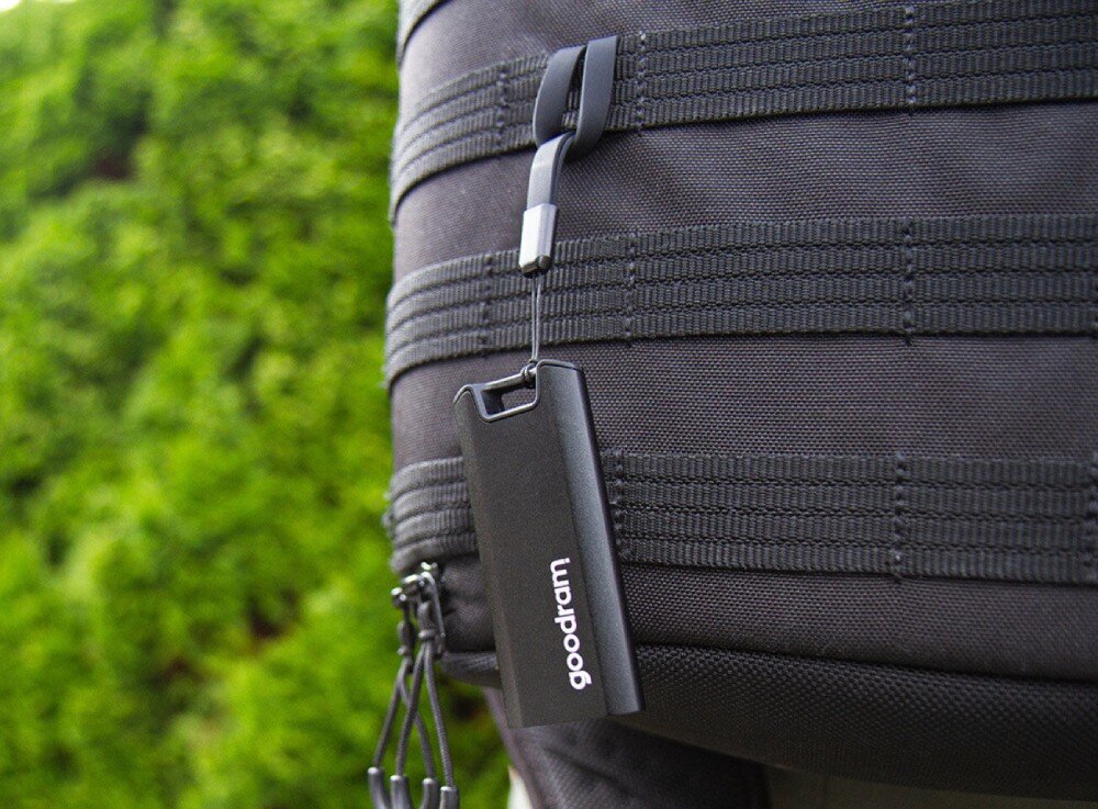 Dysk GOODRAM Move Ridge Pendrive z białym napisem 'goodram' wisi na smyczy przypiętej do poziomego pasa plecaka, w tle zielone krzewy, pojemność, kompatybilność