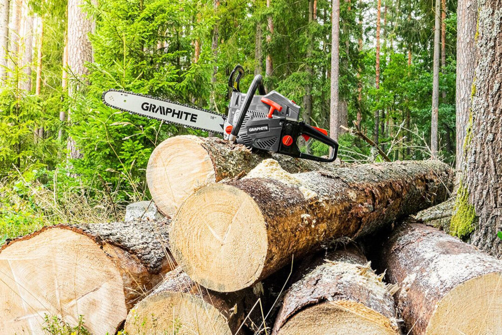 Na stosie ściętych pni drzew leży piła spalinowa z widocznym logo \'GRAPHITE\'. W tle znajduje się las z zielonymi drzewami.