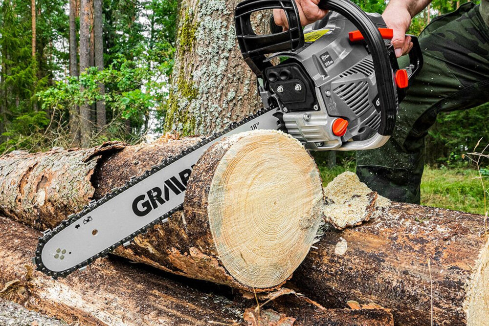 Osoba używa piły spalinowej do cięcia drewna w lesie. Na prowadnicy piły widoczny jest napis \'GRAPHITE\'.