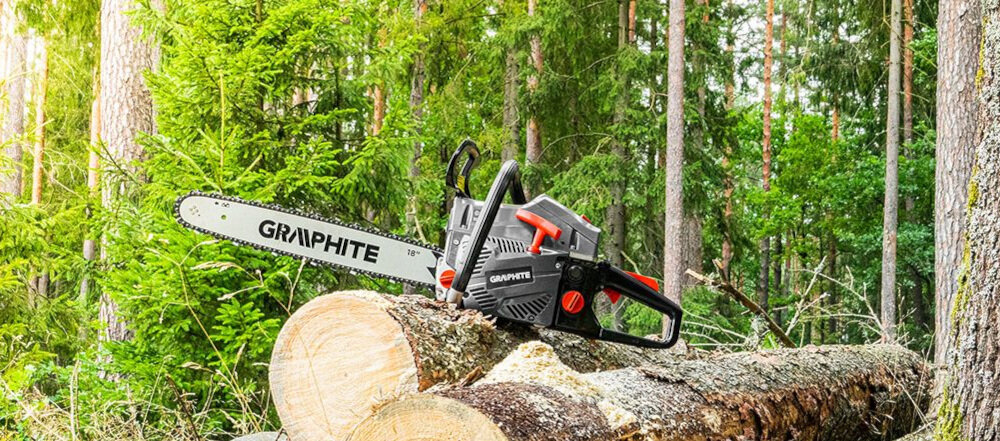Piła spalinowa z napisem \'GRAPHITE\' leży na ściętym pniu drzewa w lesie. W tle widoczne są drzewa i zielona roślinność.
