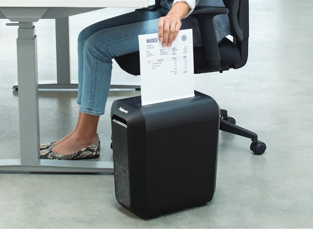 Osoba siedząca na krześle wkłada dokument z napisem \'INVOICE\' do czarnej niszczarki z widocznym logo Fellowes. Niszczarka stoi na podłodze obok biurka.