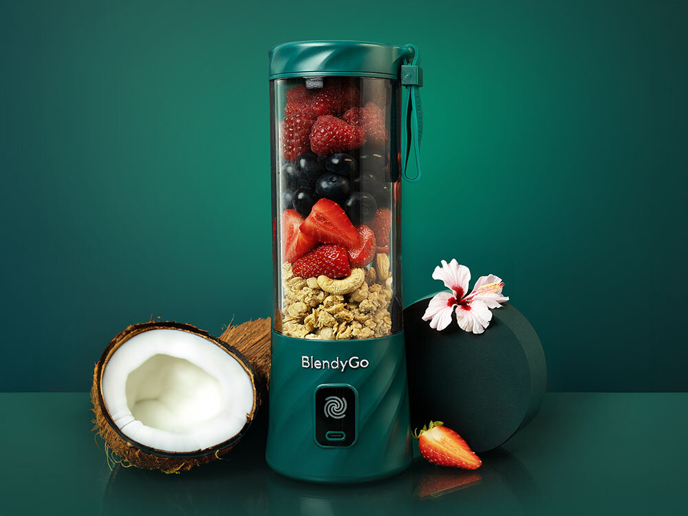 Blender personalny w kolorze butelkowej zieleni z logo \'BlendyGo\', wypełniony owocami i orzechami. Obok blendera znajduje się połówka kokosa, kwiat hibiskusa i truskawka.