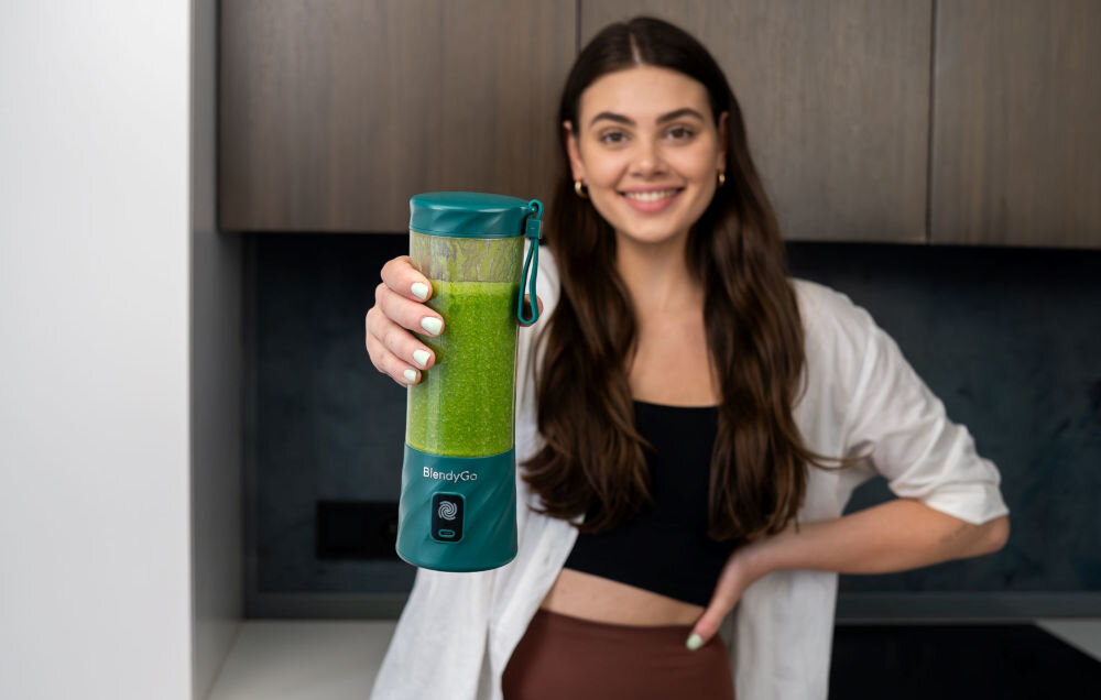 Osoba trzyma blender personalny w kolorze butelkowej zieleni z napisem \'BlendyGo\'. W tle widoczna jest kuchnia z ciemnymi szafkami.