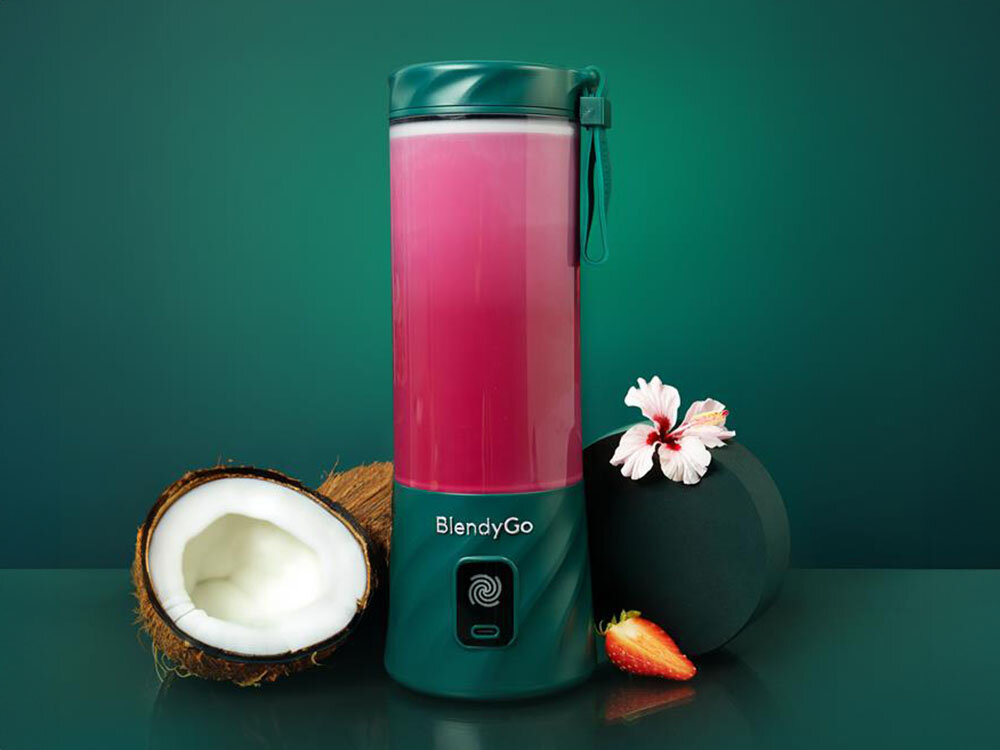 Na obrazie znajduje się blender personalny w kolorze butelkowej zieleni z napisem \'BlendyGo\'. Obok blendera leży połówka kokosa, truskawka i kwiat hibiskusa.