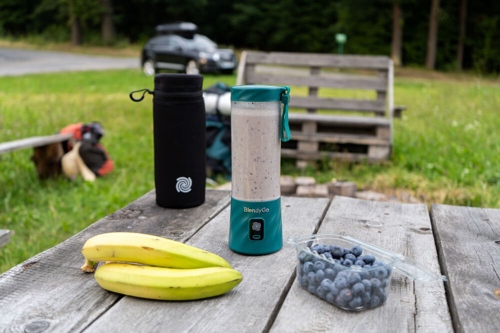 Na drewnianym stole znajduje się blender personalny BlendyGo w kolorze butelkowej zieleni, obok dwóch bananów i pojemnika z borówkami. W tle widoczny jest czarny pokrowiec z białym logo oraz samochód na trawie.
