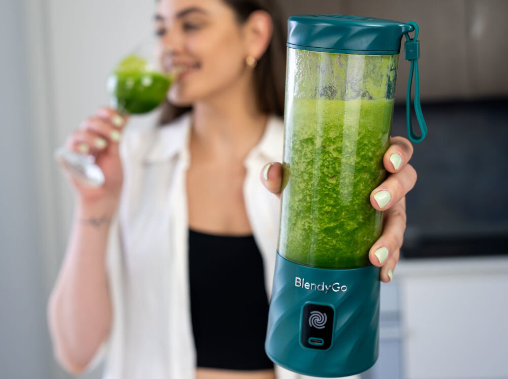 Osoba trzyma blender personalny w kolorze butelkowej zieleni z logo \'BlendyGo\', wypełniony zielonym napojem. W tle osoba pije zielony napój z kieliszka.