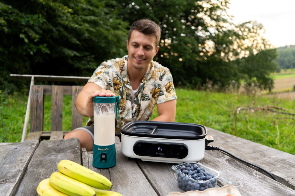 Osoba siedzi przy drewnianym stole na zewnątrz, używając blendera oznaczonego logo \'BlendyGo\'. Na stole znajdują się banany, pojemnik z borówkami i urządzenie kuchenne z wyświetlaczem.