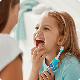 Dziecko trzyma niebieską szczoteczkę soniczną i szeroko się uśmiecha, pokazując zęby. W tle widoczna jest osoba dorosła oraz rozmyte elementy łazienki.