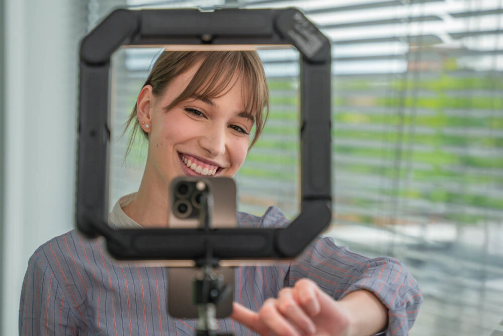 Osoba robiąca selfie za pomocą smartfona umieszczonego w kwadratowej lampie pierścieniowej. W tle widoczne są żaluzje na oknie.