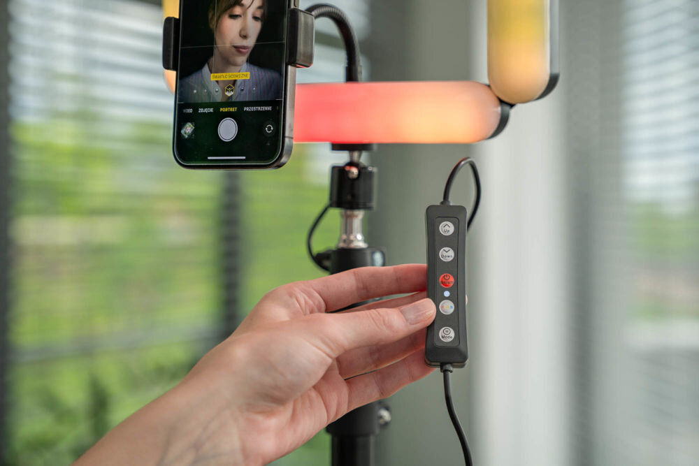 Osoba trzyma pilot do lampy pierścieniowej, na którym widoczne są przyciski sterujące. W tle znajduje się smartfon z włączoną aplikacją aparatu, a lampa świeci kolorowym światłem.
