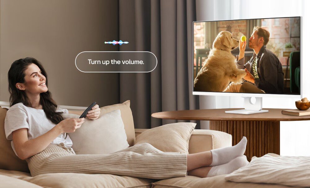 Monitor LG Smart 32U721SA-W.AEU Kobieta siedząca na kanapie, trzymająca pilota i patrząca na monitor, z dymkiem 'Turn up the volume' i falą dźwiękową, pilot, sterowanie głosem